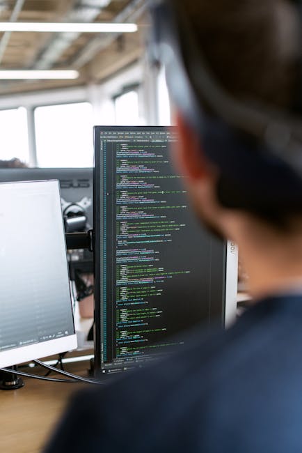 A close-up view of a person working at a computer workstation in an office environment, with their back to the camera and wearing glasses. The monitor in front displays lines of colourful code on a da