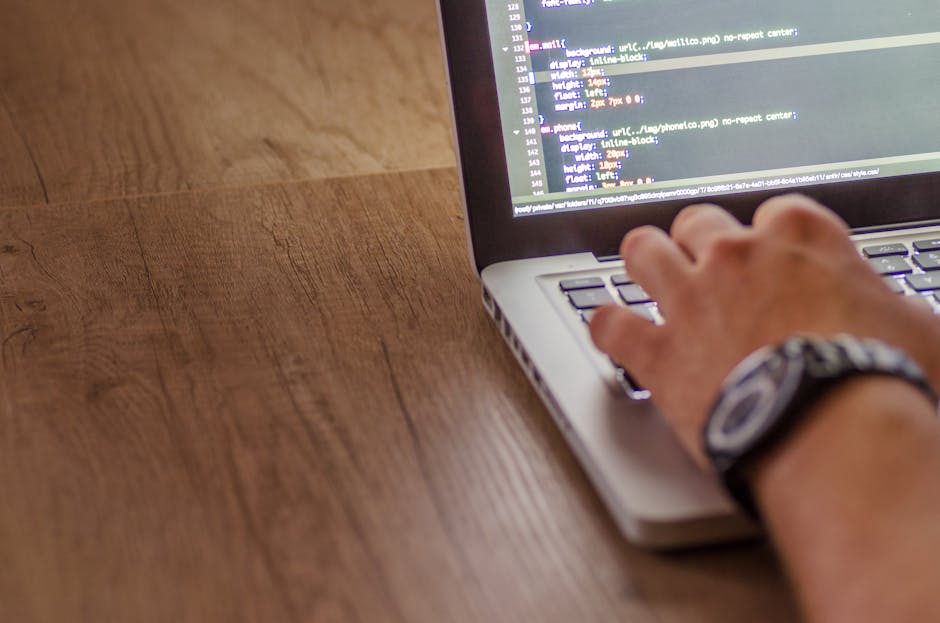 A close-up view of a person working at a computer workstation in an office environment, with their back to the camera and wearing glasses. The monitor in front displays lines of colourful code on a da